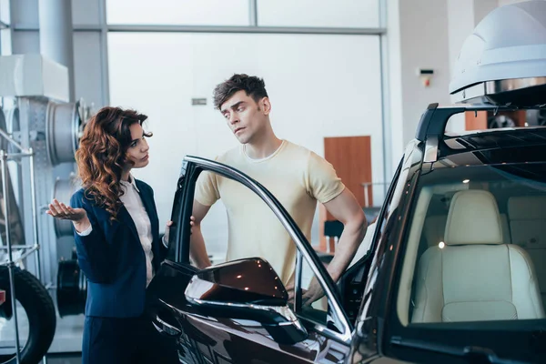 Серьезный автомобильный дилер и красивый мужчина, стоящий рядом с новой машиной — стоковое фото