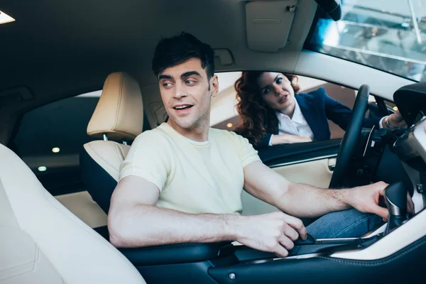 Selective focus of handsome man sitting in new car and pretty car dealer standing by car window — Stock Photo