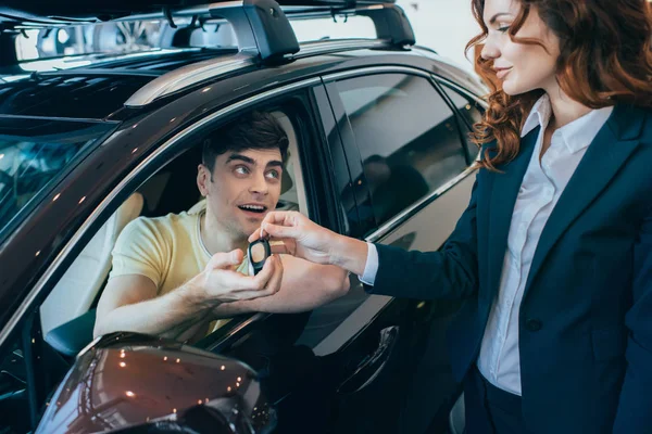 Selektiver Fokus des glücklichen Mannes, der im neuen Auto sitzt und Autoschlüssel von attraktiven Autohändlern nimmt — Stockfoto