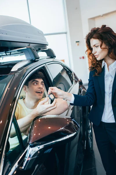 Selective focus of beautiful car dealer giving car key to happy customer sitting in new car — Stock Photo