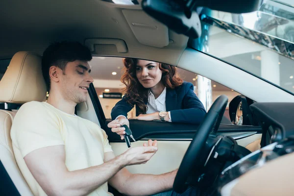 Вибірковий фокус усміхненого автодилера дає ключі щасливому чоловікові, що сидить у новому автомобілі — стокове фото