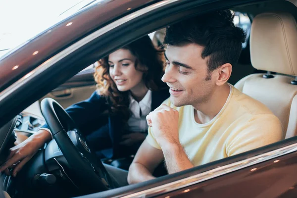 Селективный фокус улыбающегося автодилера и клиента, сидящего вместе в новом автомобиле — стоковое фото