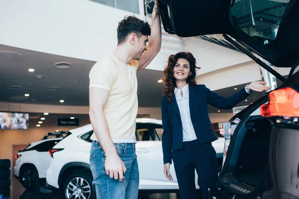 Улыбающийся автомобильный дилер и счастливый человек, стоящий рядом с новым автомобилем в автосалоне — стоковое фото