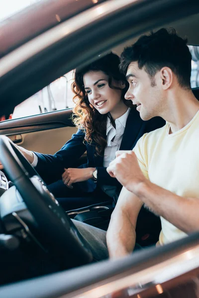 Enfoque selectivo de distribuidor de coches sonriente y hombre guapo sentado en coche nuevo juntos - foto de stock