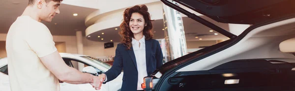 Panoramic shot of smiling car dealer and customer shaking hands in car showroom — Stock Photo