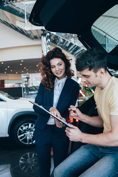Felice uomo firma carta vicino sorridente rivenditore di auto in auto showroom — Foto stock