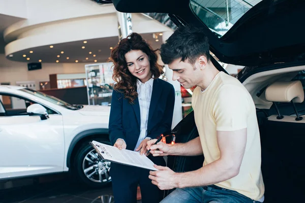 Schöner Mann signiert Dokument neben lächelndem Autohändler — Stockfoto