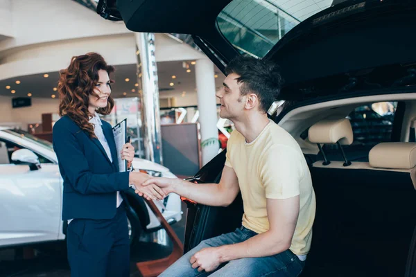 Selective focus of happy man and pretty car dealer shaking hands in car showroom — Stock Photo