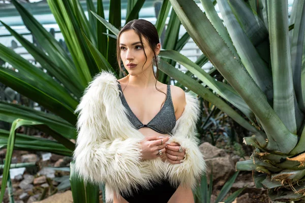 Sexy young woman in faux fur coat and underwear near plants in orangery — Stock Photo