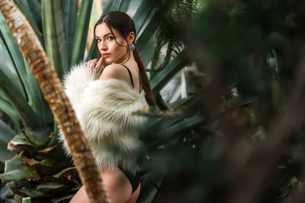 Sexy joven mujer en abrigo de piel sintética y ropa interior cerca de las plantas en el jardín botánico - foto de stock