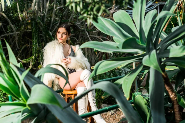 Сексуальна молода жінка в білій шубці, що сидить на стільці в ботанічному саду — стокове фото