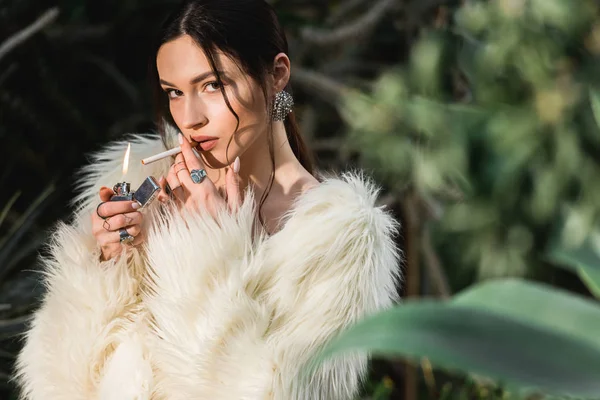 Sexy junge Frau im Kunstpelzmantel zündet sich Zigarette im botanischen Garten an — Stockfoto