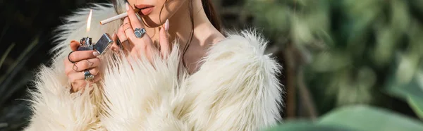 Panoramic shot of sexy young woman in faux fur coat lighting cigarette in botanical garden — Stock Photo