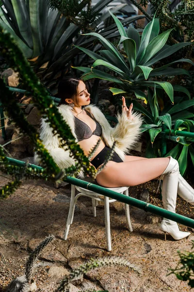 Расслабленная сексуальная женщина в искусственном шубе и нижнем белье сидит на стуле в ботаническом саду — стоковое фото