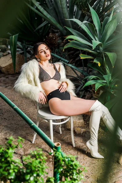Розслаблена сексуальна жінка в штучному шубі і нижній білизні сидить на стільці в ботанічному саду — стокове фото