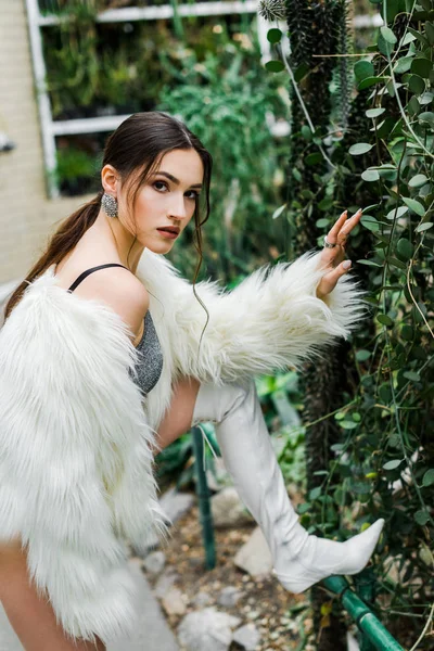 Selbstbewusstes sexy Mädchen im Kunstpelzmantel berührt grünes Laub in der Orangerie — Stockfoto