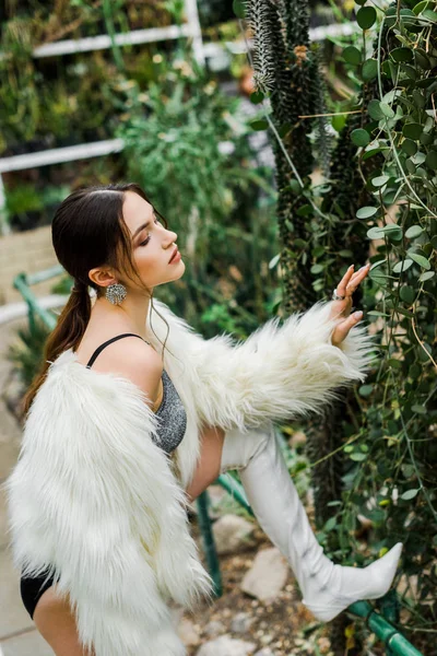 Confident sexy girl in faux fur coat touching green foliage in orangery — Stock Photo