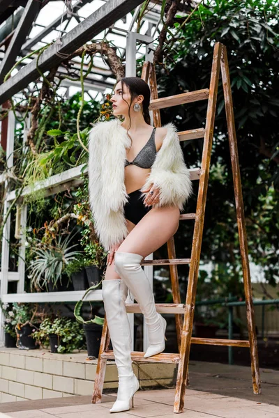 Sexy pensive girl in faux fur coat and underwear standing near wooden ladder in orangery — Stock Photo