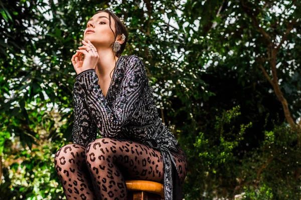 Low angle view of sexy pensive woman in black pantyhose sitting on chair in botanical garden — Stock Photo