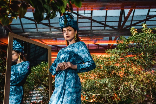 Beautiful woman in blue turban and dress standing near mirror in orangery — Stock Photo