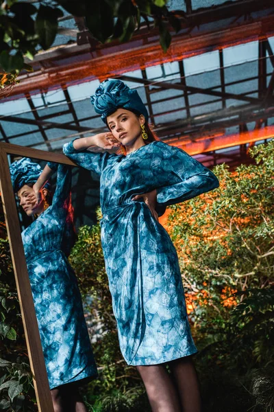 Красивая женщина в голубом тюрбане и платье, стоящее возле зеркала в оранжерее — стоковое фото