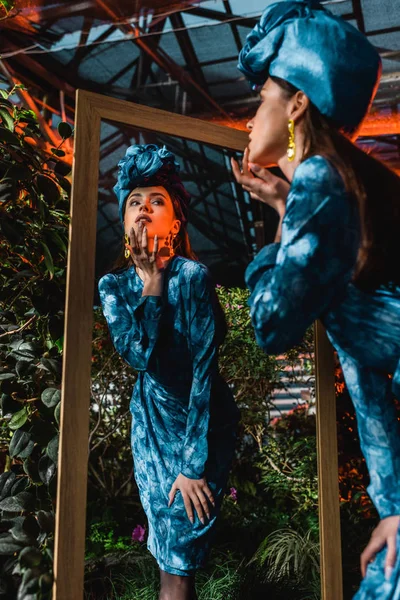 Tiefansicht der charmanten Frau in blauem Kleid und Turban, die in der Orangerie in den Spiegel schaut — Stockfoto