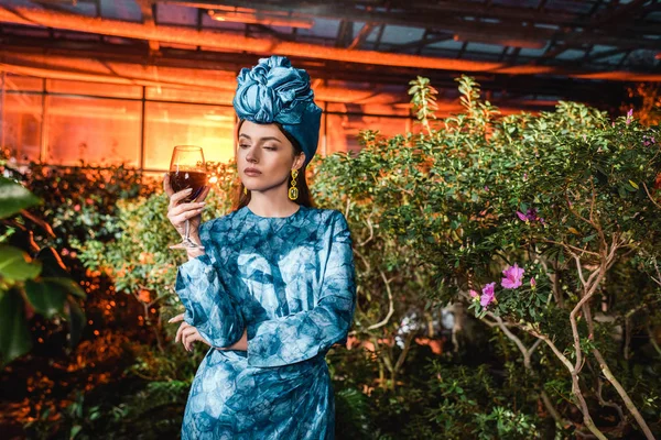 Mulher atraente em turbante azul segurando copo de vinho tinto em orangery — Fotografia de Stock