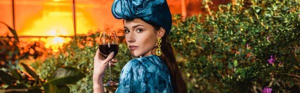 Tiro panorâmico de mulher atraente em turbante azul segurando copo de vinho tinto em orangery — Fotografia de Stock