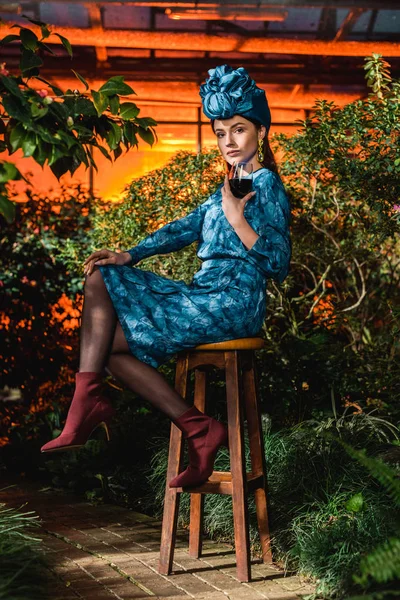 Mulher bonita em vestido e turbante sentado na cadeira e segurando copo de vinho em orangery — Fotografia de Stock