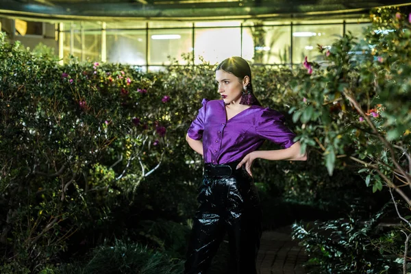 Elegante junge Frau in lila Bluse steht mit Armen akimbo in Orangerie — Stockfoto