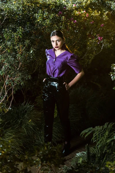 Elegante junge Frau in lila Bluse steht mit Armen akimbo in Orangerie — Stockfoto