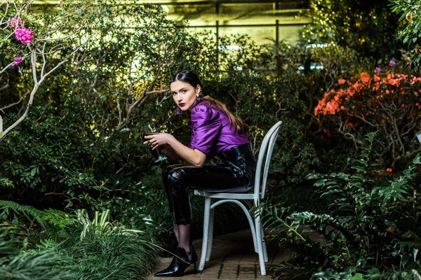 Attraktive Frau in lila Bluse und Lederhose sitzt auf Stuhl und hält Weinglas in der Orangerie — Stockfoto