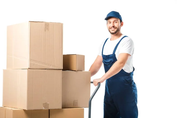 Handsome mover in uniform looking at camera while transporting cardboard boxes on hand truck isolated on white — Stock Photo