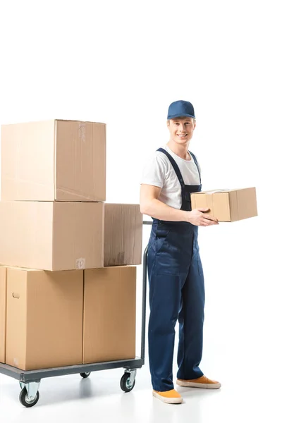 Sorrindo bonito mover em uniforme transportando caixa de papelão perto de caminhão de mão com pacotes isolados em branco — Fotografia de Stock