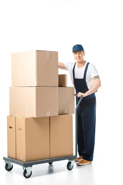 Bonito mover no uniforme sorrindo ao transportar caixas de papelão no caminhão de mão isolado no branco — Fotografia de Stock