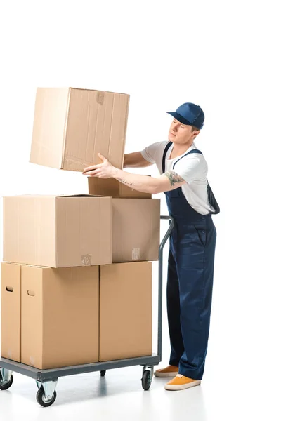 Handsome mover in uniform transporting cardboard box near hand truck with packages isolated on white — Stock Photo