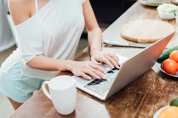 Vista parcial da mulher jovem digitando no teclado do laptop na cozinha — Fotografia de Stock