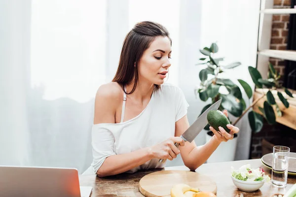 Sexy Mädchen schneidet Avocado mit Messer an Holztisch in Küche — Stockfoto