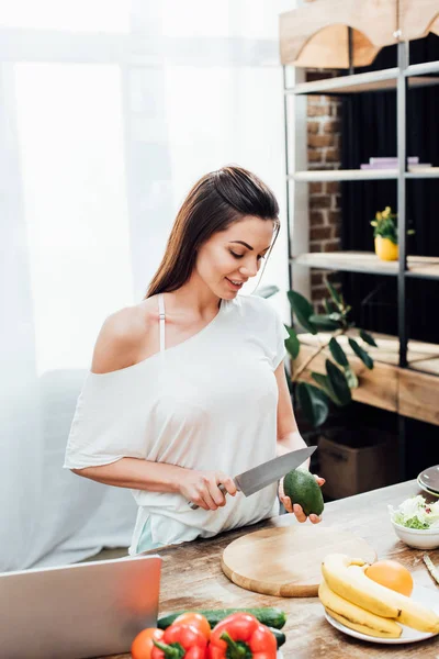 Lächelnde junge Frau schneidet Avocado an Holztisch in Küche — Stockfoto