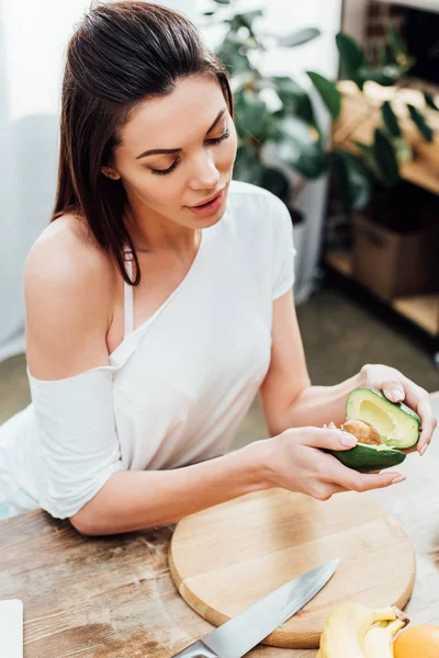 High angle view of pretty stylish girl holding cut avocado near table in kitchen — Stock Photo