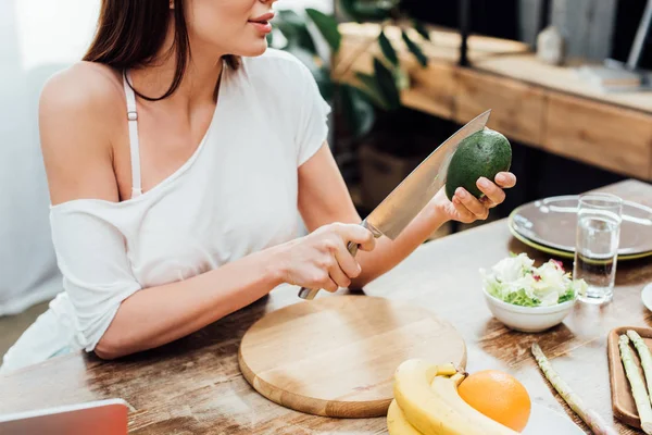 Vista ritagliata di ragazza taglio avocado con coltello al tavolo di legno in cucina — Foto stock