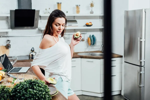 Menina muito elegante segurando cortar abacate perto da mesa na cozinha — Fotografia de Stock