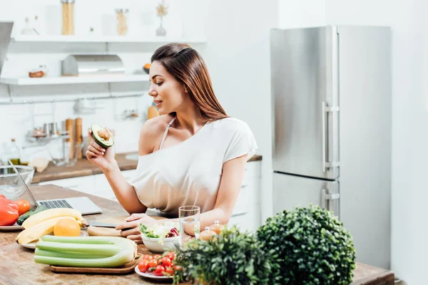 Досить стильна дівчина тримає вирізане авокадо біля столу на кухні — стокове фото