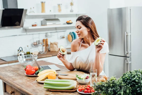 Menina muito elegante segurando cortar abacate perto da mesa na cozinha — Fotografia de Stock