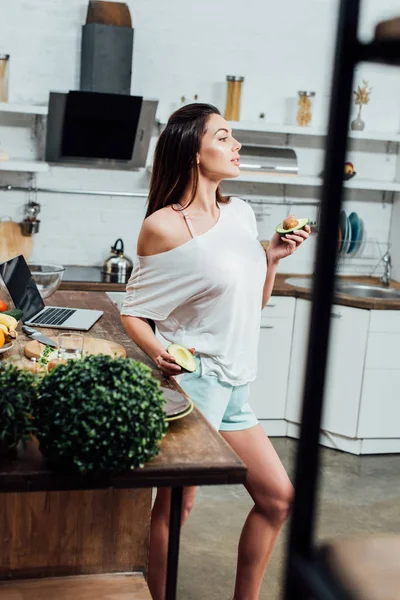 Pretty stylish girl holding cut avocado near table in kitchen — Stock Photo