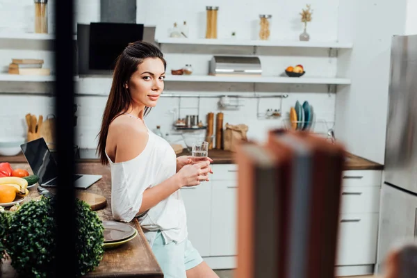 Attractive girl standing near wooden table and holding glass of water in kitchen — Stock Photo