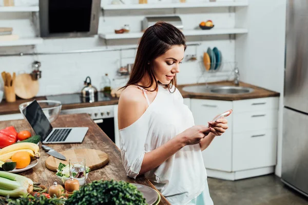 Menina atraente usando smartphone perto de mesa de madeira na cozinha — Fotografia de Stock