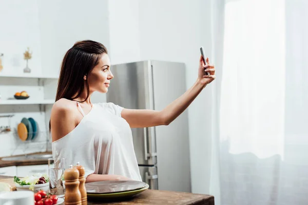 Jovem encantadora tomando selfie perto da mesa na cozinha — Fotografia de Stock