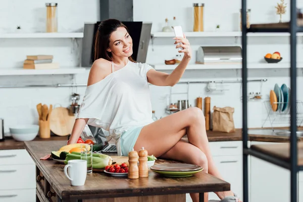 Smiling girl sitting on wooden table and taking selfie in kitchen — Stock Photo