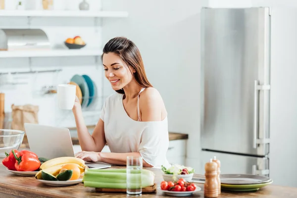 Menina encantadora beber café ao usar laptop na cozinha — Fotografia de Stock